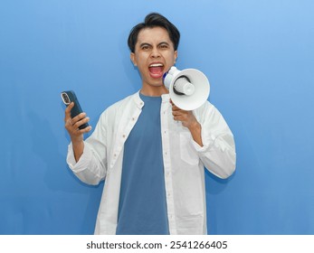 Young Asian man with an angry expression, holding a megaphone and using a mobile phone on a blue background. Ideal for communication or announcement concepts.