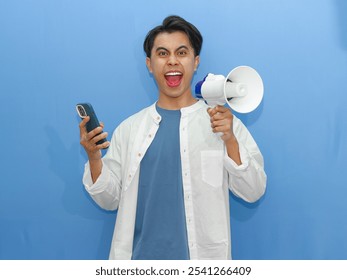 Young Asian man with an angry expression, holding a megaphone and using a mobile phone on a blue background. Ideal for communication or announcement concepts.