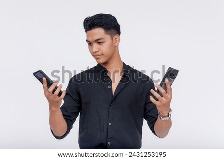 A young Asian man appears indecisive while comparing two smartphones, isolated on a clean white background. Cellphone shopping and specification comparison concepts.