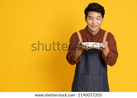 Young Asian man in apron standing and holding empty white plate or dish isolated on yellow background