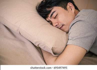 Young Asian Man With Attractive Smile Sleeping And Laying On Bed Indoors. Portrait Of Asian Man Sleep Tight In The Bedroom