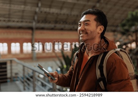 Young asian man with backpack using smart phone app, smiling and looking away in train station