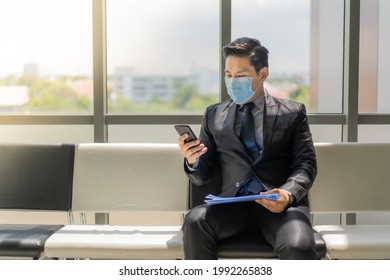 Young Asian Man In Black Suit And Tie, Wearing A Face Mask For Protection Covid-19 Sitting On Chair In Office Lobby, Holding A Blue Folder And Looking At His Phone While Waiting For A Job Interview.