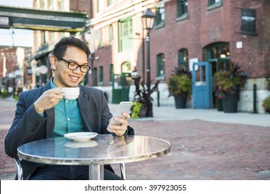 Young asian man in business casual attire sitting and smiling in relaxing outdoor cafe drinking cup of coffee while using mobile phone, with copy space.