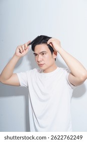 Un joven asiático se cepilla cuidadosamente el cabello con un peine mientras está de pie sobre un fondo claro. Parece centrado en lograr un peinado ordenado.
