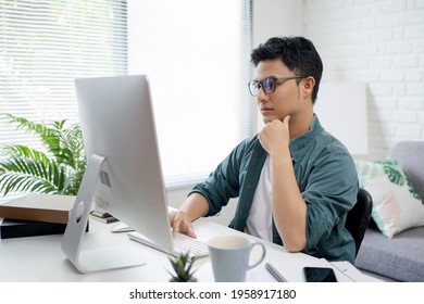 Young Asian man in casual clothes is sitting and using a laptop on the desk in the room. He works very seriously. - Powered by Shutterstock