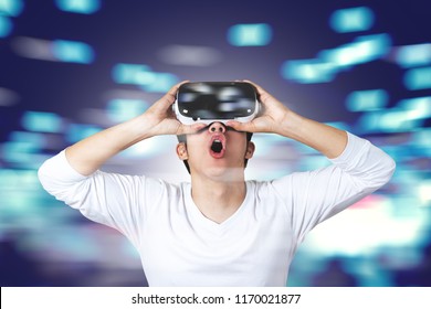 Young Asian Man In Casual White Wearing And Holding VR Glasses By Hands Looking Up Above Feeling Excited And Amazed On Blue Motion Technology Blurred Background. Virtual Reality Experience Concept.