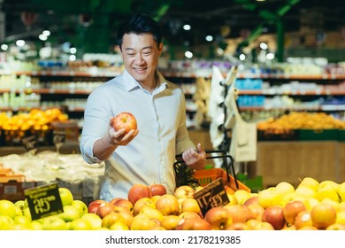 young asian man chooses and picks in eco bag apple fruit or vegetables in the supermarket. male customer standing a grocery store near the counter buys and throws in a reusable package in market - Powered by Shutterstock