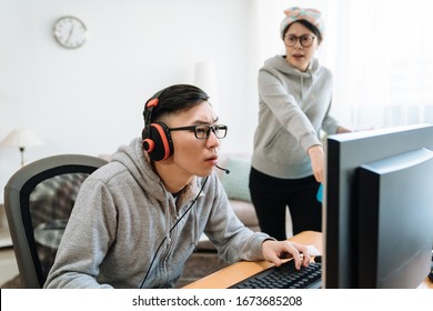 Young Asian Man Concentrated Wear Headphones And Staring At Computer Screen Playing Video Online Game. Angry Girlfriend Scolding Her Lazy Boyfriend. Lady Clean Up Messy Room And Getting Mad At Home