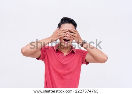 A young asian man covers his face but peeping with one eye. Too impatient to wait for the surprise. Isolated on a white background. Spoiler alert concept.