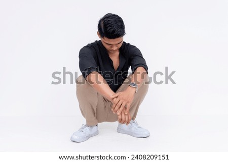 A young asian man crouching down and looking to the ground, looking depressed. Studio shot isolated on a white background.