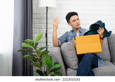 Young Asian Man Dissatisfied Opening Parcel At Home, Sitting On Couch With Cardboard Box, Angry Displeased Customer Confused By Wrong Or Damaged Order, Bad Delivery Shipping Service Concept