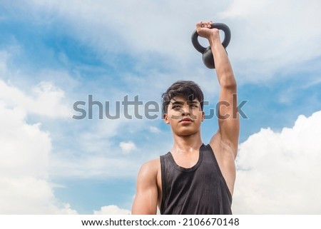 A young asian man does Kettlebell presses outside during a clear day. Outdoor shoulder workout and training.