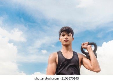 A Young Asian Man Does One Arm Kettlebell Presses Outside During A Clear Day. Outdoor Shoulder Workout And Training.