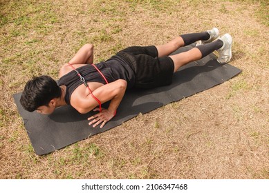 A young asian man does a set of pushups with resistance bands on a black mat at an open field outdoors. Chest or full body workout.