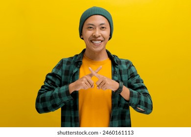 A young Asian man, dressed in a beanie hat and a casual shirt, is making an X sign gesture by crossing his fingers while standing against a yellow background - Powered by Shutterstock