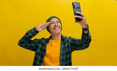 A young Asian man, dressed in a beanie hat and casual clothes, salutes while taking a selfie with his smartphone against a yellow background - Powered by Shutterstock