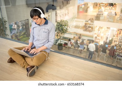 Young Asian Man Dressed In Casual Style Working With His Laptop While Listening To Music. Digital Nomad Working In Co Working Space, Modern IT Lifestyle With Work Life Balance Concept.