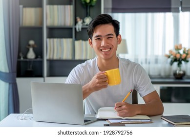 Young Asian Man Drinking Coffee And Take Notes, Work At Home, Looking At Camera.