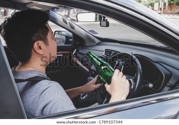 Young asian man drives a car with drunk a\
bottle of beer behind the wheel of a\
car.
