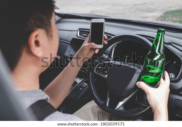 Young asian man\
drives a car with drunk a bottle of beer and using smartphone\
behind the wheel of a\
car.