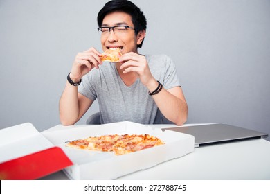 Young Asian Man Eating Pizza In Office Over Gray Background