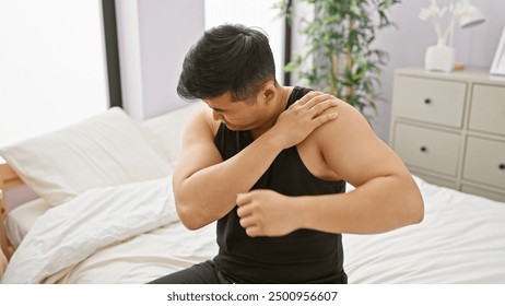 Young asian man experiencing shoulder pain in his bedroom, showing discomfort with a grimace. - Powered by Shutterstock