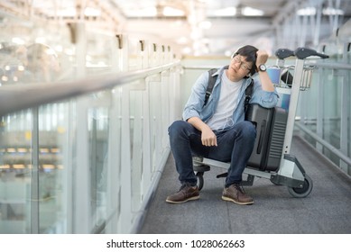 Young Asian Man Feeling Exhausted Sitting On Airport Trolley With His Suitcase Luggage In The International Airport Terminal, Flight Problem And Travel Insurance Concepts