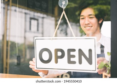 Young Asian Man Flip The Open Label To Welcome Customer And Open The Coffee Shop In Morning. Male Asian Barista Open The Coffee Cafe At Doorway With Reflection In Mirror.