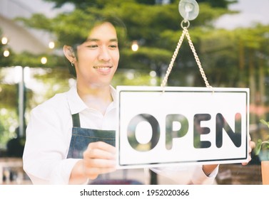 Young Asian Man Flip The Open Label To Welcome Customer And Open The Coffee Shop In Morning. Male Asian Barista Open The Coffee Cafe At Doorway With Reflection In Mirror.