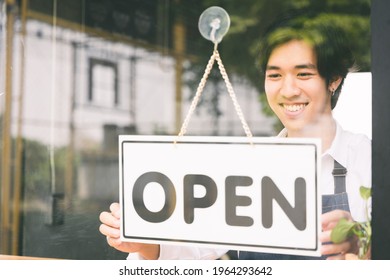 Young Asian Man Flip The Open Label To Welcome Customer And Open The Coffee Shop In Morning. Male Asian Barista Open The Coffee Cafe At Doorway With Reflection In Mirror.