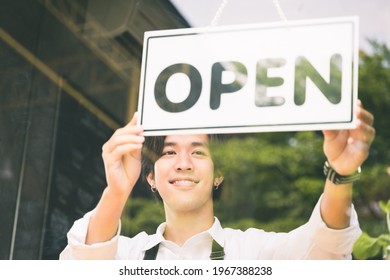 Young Asian Man Flip The Open Label To Welcome Customer And Open The Coffee Shop In Morning. Male Asian Barista Open The Coffee Cafe At Doorway With Reflection In Mirror.