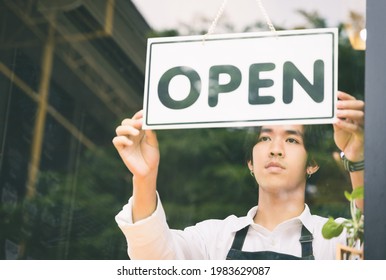 Young Asian Man Flip The Open Label To Welcome Customer And Open The Coffee Shop In Morning. Male Asian Barista Open The Coffee Cafe At Doorway With Reflection In Mirror.