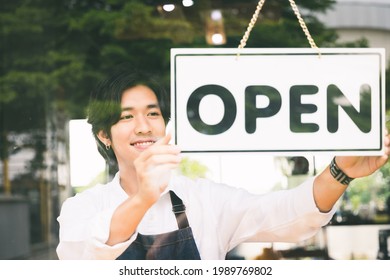 Young Asian Man Flip The Open Label To Welcome Customer And Open The Coffee Shop In Morning. Male Asian Barista Open The Coffee Cafe At Doorway With Reflection In Mirror.