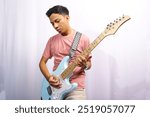 a young asian man focused on playing a light blue electric guitar with a serious facial expression and a checkered guitar strap. wearing a pink t-shirt and a white background.