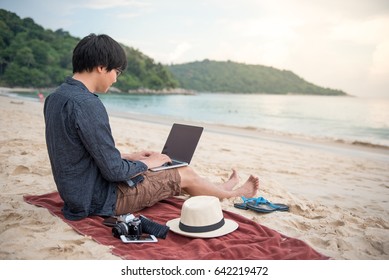 Young Asian Man Freelancer Working With Laptop Computer On Tropical Island Beach. Digital Nomad Life Of Freelance Lifestyle On Summer Holiday Vacation. Remote Work And Freedom Travel Concept