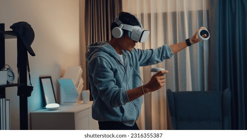 Young Asian man gamer wear gray hoodie with VR oculus hold VR joystick controllers feeling exciting with new experience of game stand front sofa living room home at night. Virtual reality concept. - Powered by Shutterstock