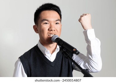 A young Asian man is giving a speech into a microphone with his fist in the air, symbolizing strength. He is delivering a political message. - Powered by Shutterstock