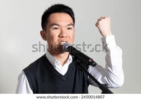 A young Asian man is giving a speech into a microphone with his fist in the air, symbolizing strength. He is delivering a political message.