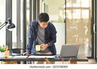 Young Asian Man Graphic Designer Standing At Office Desk And Using Laptop Computer.