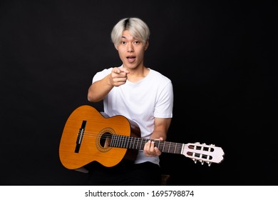 Young Asian Man With Guitar Over Black Background Surprised And Pointing Front