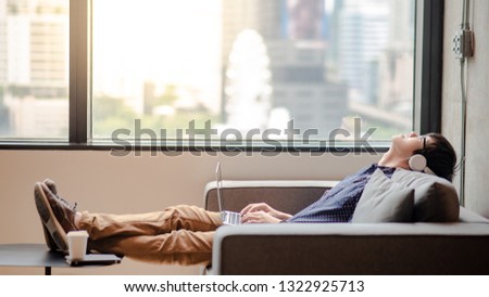 Young Asian man with headphones and eyeglasses lying down on confortable sofa while enjoy listening to music from laptop computer. Urban lifestyle in living space. Relaxation concept