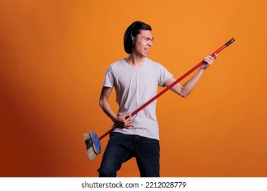 Young asian man in headphones listening to music, playing virtual guitar, imagining rockstar lifestyle. Musician practicing, teenager using mop as musical instrument, fun pastime, leisure activity