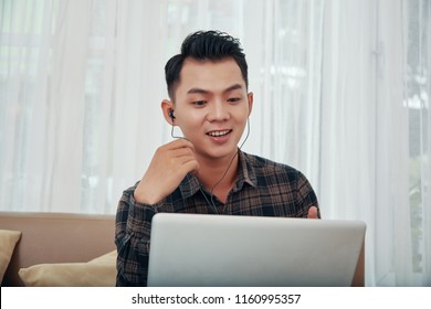 Young Asian Man In Headphones Speaking Via Laptop Having Video Call While Sitting On Sofa At Home 