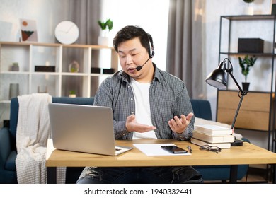 Young Asian Man In Headset Sitting At Desk With Laptop And Talking During Video Call. Businessman Using Modern Devices For Online Conversation, Working Online From Home Office