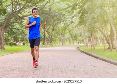 Young Asian man, healthy looking, wearing blue tank top, jogging on concrete road in park, with many trees and green lawn as background, in Thailand. - Powered by Shutterstock