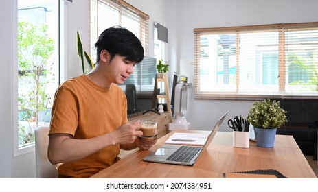 Young Asian Man Holding Coffee Cup And Reading News On Laptop Computer.