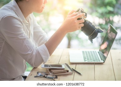 Young Asian man holding digital single-lens reflex camera (DSLR) checking photo on camera screen display sitting with laptop computer and smartphone on the table. Photography, art and hobby concepts - Powered by Shutterstock