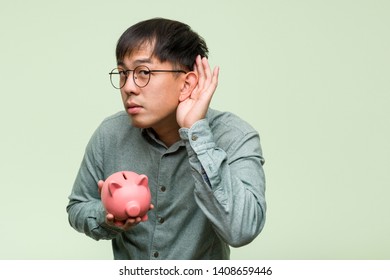 Young asian man holding a piggy bank try to listening a gossip