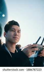 Young Asian Man Holding  Radio Remote Controller On The Street. Boy Flying A Drone Outside At Daytime. Quadcopter Operator Standing In Sunlight. Shallow Depth Of Field.